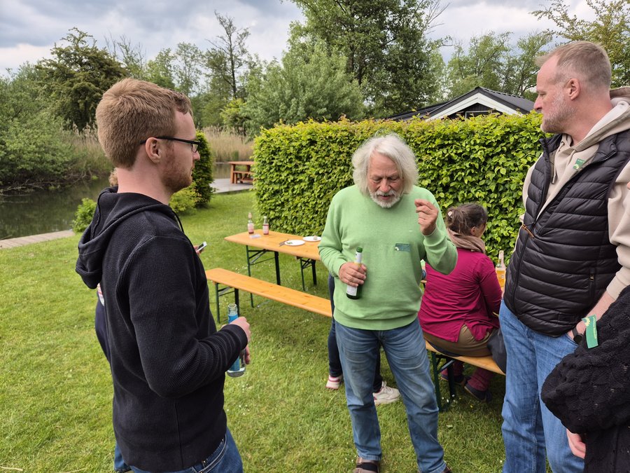 Im Vordergrund unterhalten sich 3 Personen, die auf einer Wiese stehen. Links im Hintergrund fließt ein Fluss, auf der Wiese stehen Biertischgarnituren, auf denen Flaschen sowie Teller und Besteck stehen und liegen. Im Hintergrund eine grüne Hecke sowie Personen, die an den Biertischen sitzen.